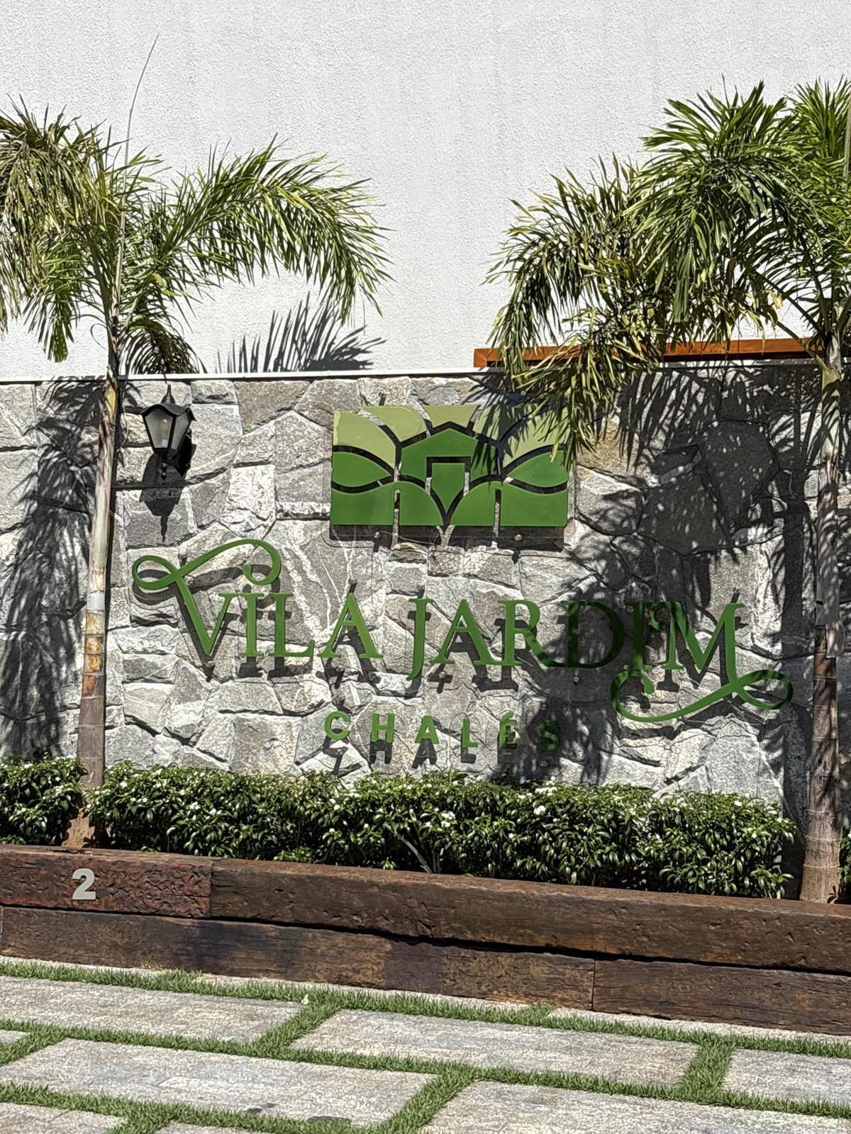 Placa da Vila Jardim Chalés em muro de pedra, cercada por palmeiras